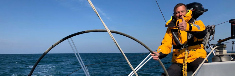 Photo à bord d'un voilier d'une personne en veste de quart orange tenant la barre regardant l'objectif avec un pouce levé