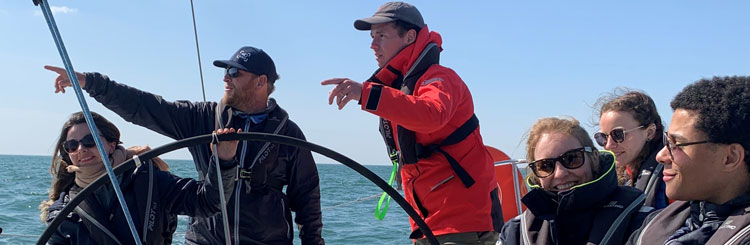 Photo prise dans un bateau d'un groupe de personne à assises à côté de la barre. Deux personnes debouts pointent l'horizon et une personne tient la barre.